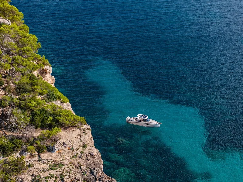 NAVAN S30 anchored alone beneath a steep pine-covered cliff on Ibiza, where deep blue water meets a turquoise shallow shelf