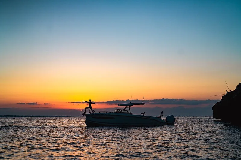 Silueta del NAVAN S30 fondeado junto a un cabo de Ibiza al atardecer, con una invitada de pie en la proa contra el cielo naranja