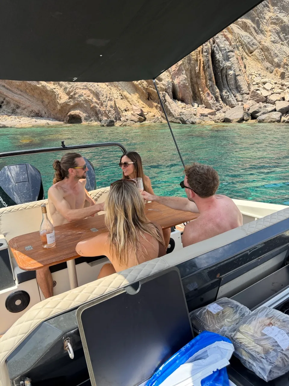 Vier collega's delen een fles rosé aan de teakhouten tafel van de NAVAN S30 voor anker in een rotsbaai