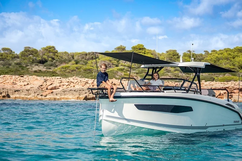 Two guests relaxing on the bow of the NAVAN S30 beneath the shade sails, with a rocky pine-covered shoreline behind